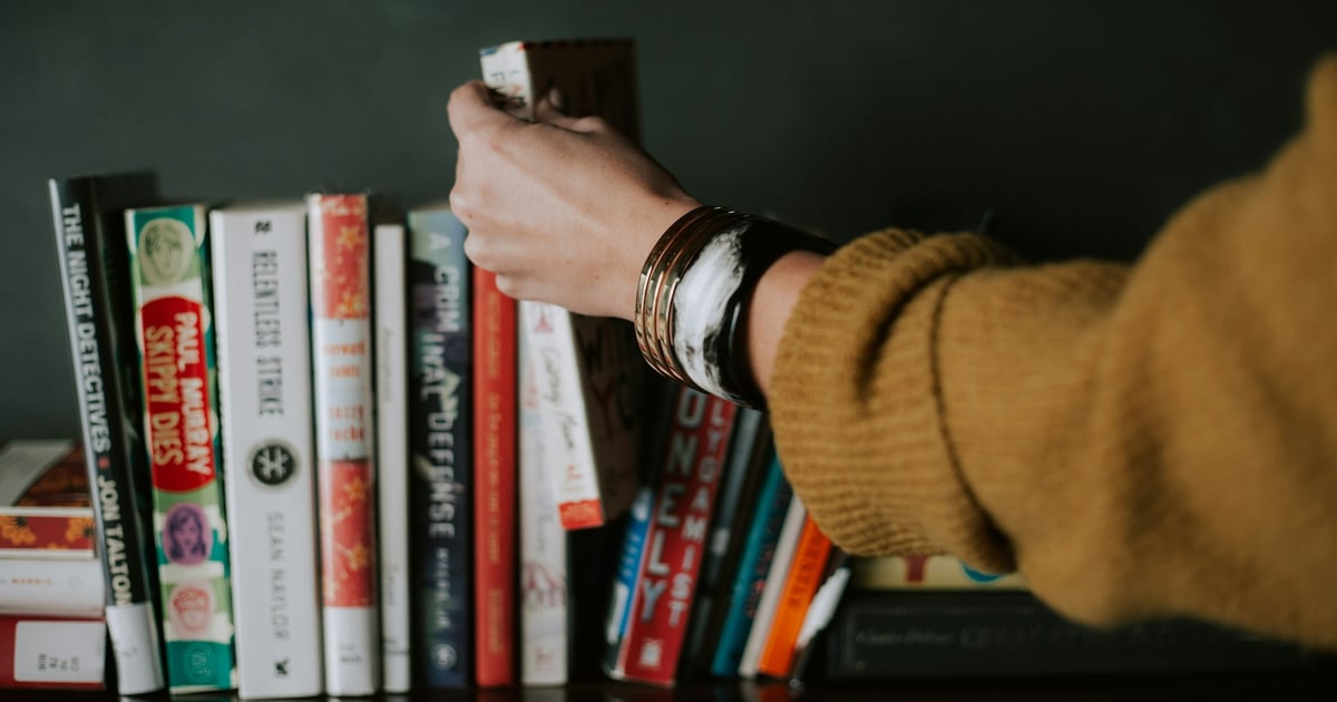 A person wearing a mustard-colored sweater is reaching for a book on a shelf filled with hardcover books.